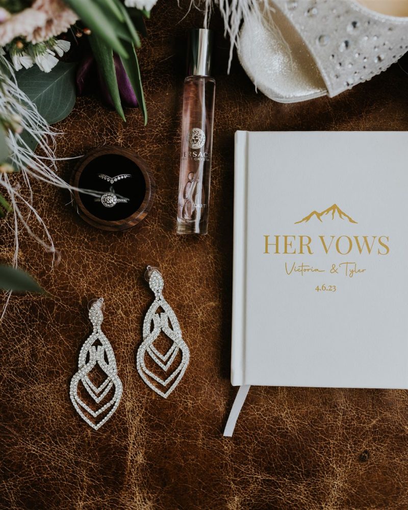A flat lay of wedding items on a brown leather surface, perfect for a photographer: a Her Vows book, crystal earrings, perfume bottle, ring box, white heels, and a floral bouquet—ideal inspiration for your elopement day.