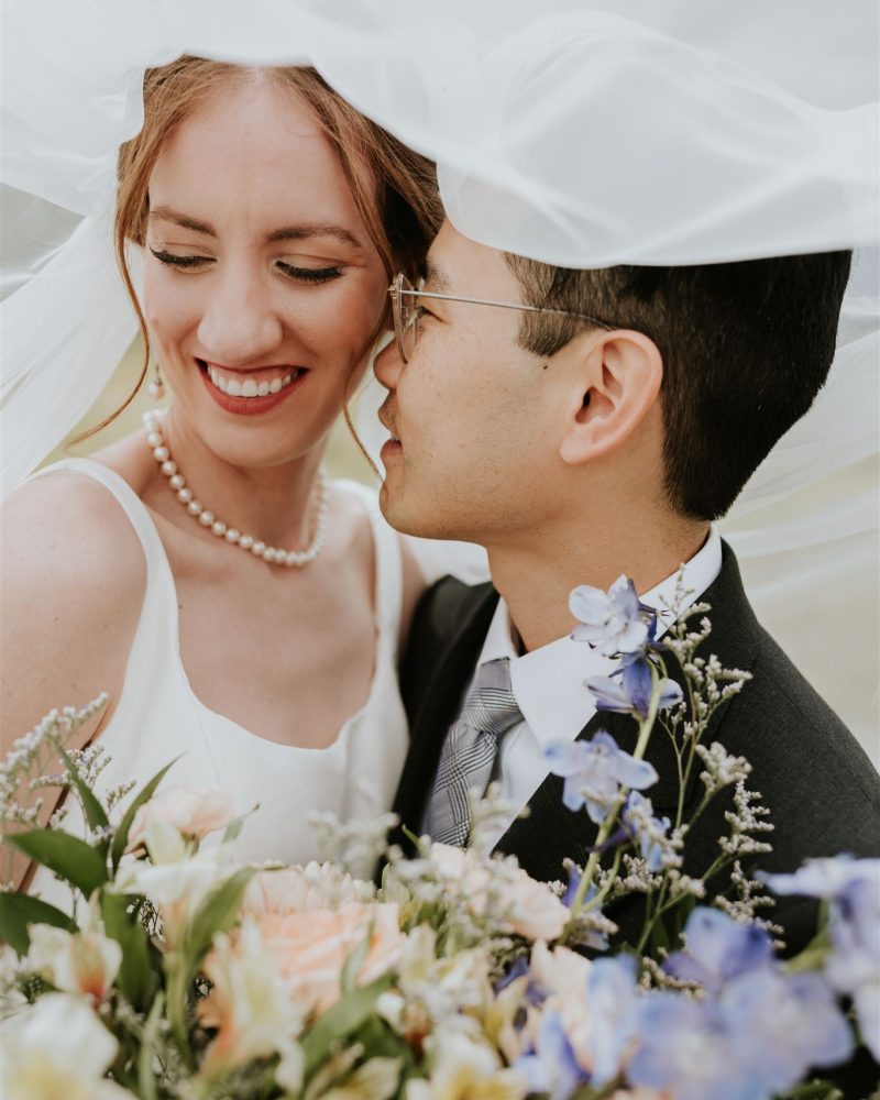 A bride and groom stand close together under a veil, smiling joyfully. Captured by a Canmore photographer, the bride holds a bouquet of pastel flowers, and both appear happy and in love on their wedding day.