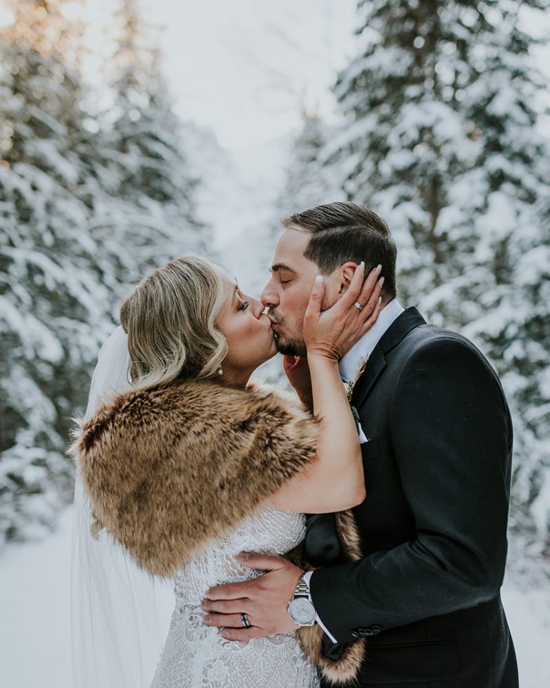 Bride and groom share a kiss in a snowy Canmore forest, surrounded by snow-covered trees. Perfect for an intimate elopement, the bride wears a white dress, veil, and brown fur shawl, while the groom is in a black suit.
