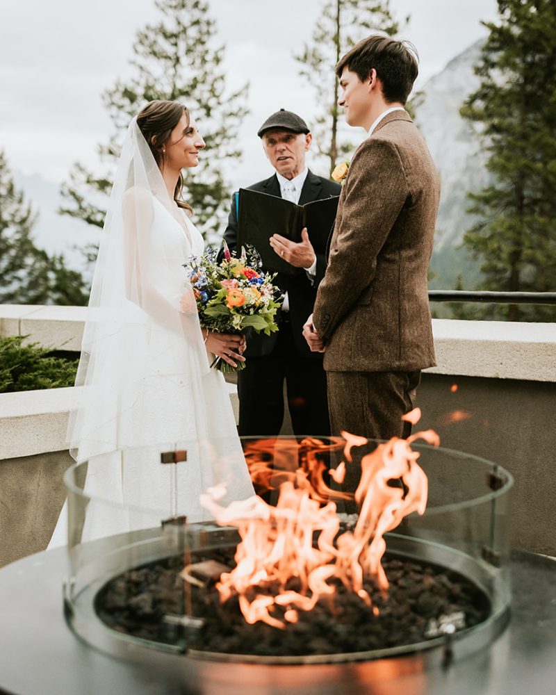 A bride and groom stand facing each other during a Calgary wedding ceremony, with an officiant between them. A glass fire pit burns in the foreground, framed by trees and majestic mountains in the background.