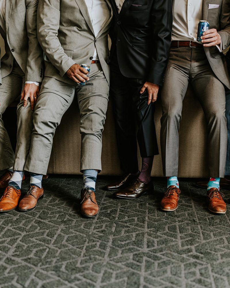 A group of men in suits stand side by side, showing off their colorful, patterned socks and brown dress shoes as they slightly lift or pull up their pant legs during a Calgary elopement.