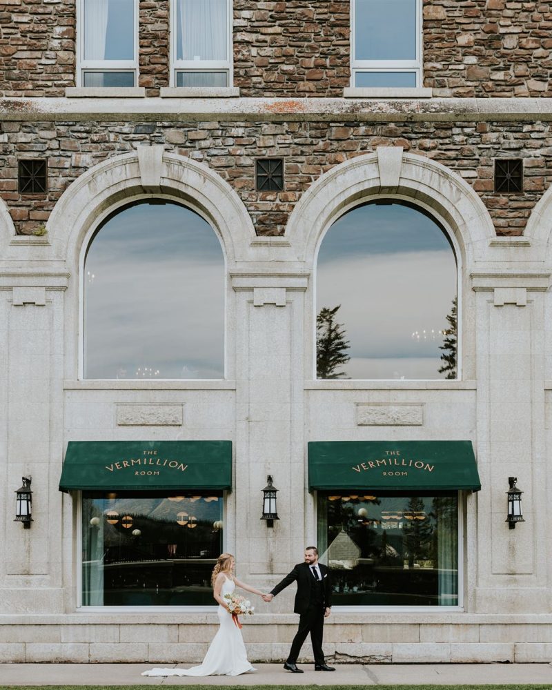 A bride and groom hold hands and smile at each other outside a stone building with large arched windows and green awnings that read VERMILLION ROOM, beautifully captured by a Canmore photographer. Trees and mountains are reflected in the windows.