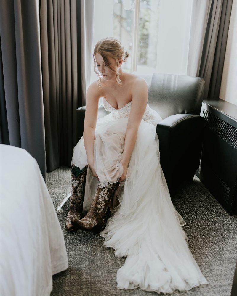 A bride in a white wedding dress sits on a black chair by a window, putting on ornate cowboy boots. Light streams in from behind, illuminating her and the flowing gown—capturing the intimate feel of a Banff elopement.