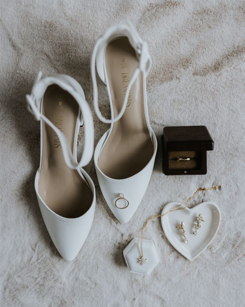 A pair of white heels, a gold ring, a necklace, earrings, and two rings in a dark box are arranged on soft, light-colored fabric—perfect details for a Banff wedding photographer to capture.