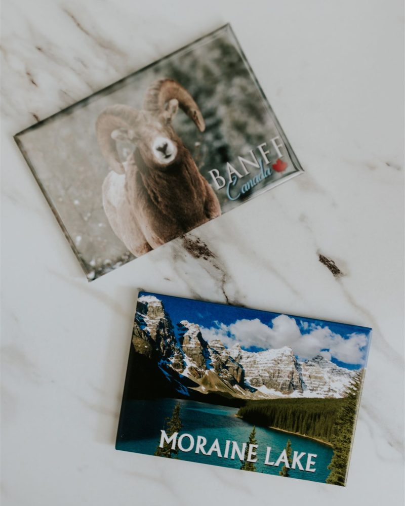 Two souvenir magnets on a marble surface: one with a ram and the word BANFF, and one with a scenic mountain lake and the words MORAINE LAKE—perfect inspiration for a wedding or elopement photographer seeking breathtaking destinations.