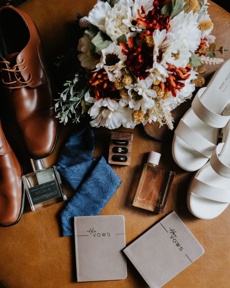 A flat lay of wedding items on a brown surface, perfect for a Canmore elopement, featuring a bouquet of flowers, brown dress shoes, white heeled sandals, blue tie, perfume bottles, earrings, and His Vows and Her Vows notebooks captured by a photographer.