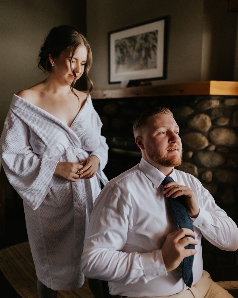 A woman in a robe smiles while standing behind a man seated on a chair, adjusting his tie. They appear to be getting ready for an intimate elopement in a cozy room with a stone fireplace and framed picture, perhaps near Banff.