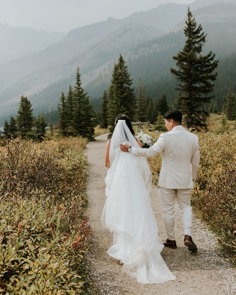 A bride and groom walk hand in hand down a scenic path surrounded by greenery and pine trees in Banff, with mountains visible in the background. Captured by a photographer, their elopement is framed by nature’s beauty.
