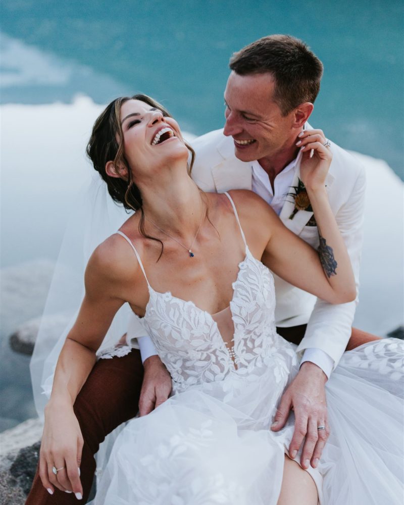 A joyful bride and groom sit together by a turquoise lake, laughing and smiling. Captured by a Calgary wedding photographer, the bride in a white lace dress and veil, and the groom in a white suit jacket, hold each other lovingly.