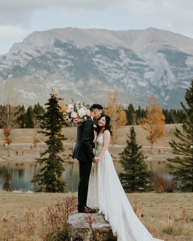 A bride and groom stand on a rock in an outdoor setting near Canmore, holding hands and smiling. Behind them are autumn trees, a tranquil lake, and a large mountain under a cloudy sky—perfect for an intimate elopement.