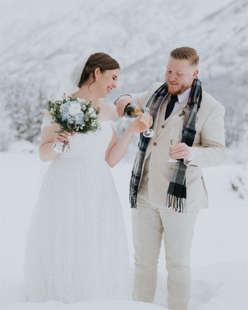 A bride in a white dress and bouquet smiles as a groom in a scarf and light suit pours champagne into her glass. Their wedding is captured by a photographer amid Banff’s snowy, mountainous landscape.