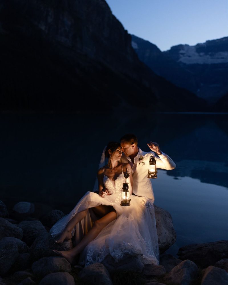 A bride and groom sit on rocks by a lake at dusk near Canmore, holding glowing lanterns. Mountains and water are visible in the background, creating a romantic and serene elopement atmosphere.