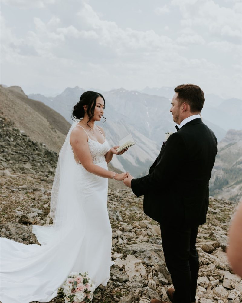 A bride and groom stand on a rocky mountain top near Calgary, holding hands as the bride reads vows from a book. She wears a white dress and veil; he’s in a black suit. Their wedding bouquet rests beside them, framed by stunning mountains.