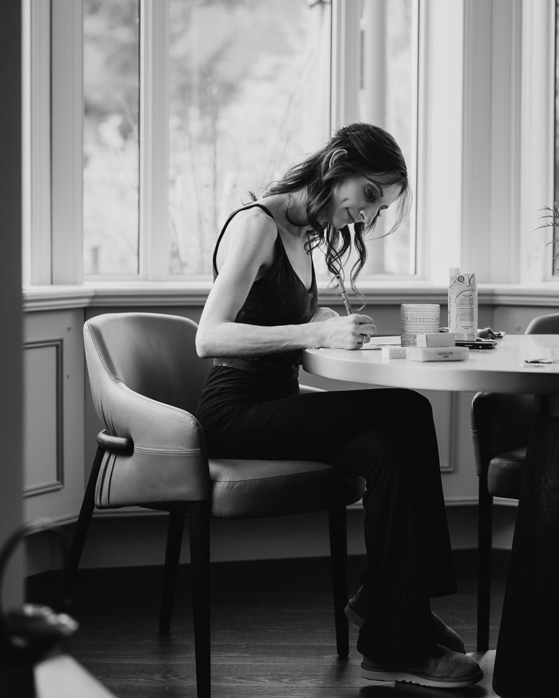 A woman sits at a round table by a large window in Calgary, focused on writing or drawing. She is dressed in dark clothing, bathed in natural light. The black and white scene captures a quiet moment before a wedding.
