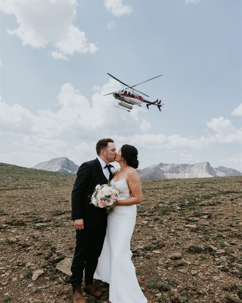 A bride and groom share a kiss outdoors on rocky terrain in Banff, mountains rising behind them; a helicopter flies overhead in the partly cloudy sky as their photographer captures this unforgettable elopement moment.