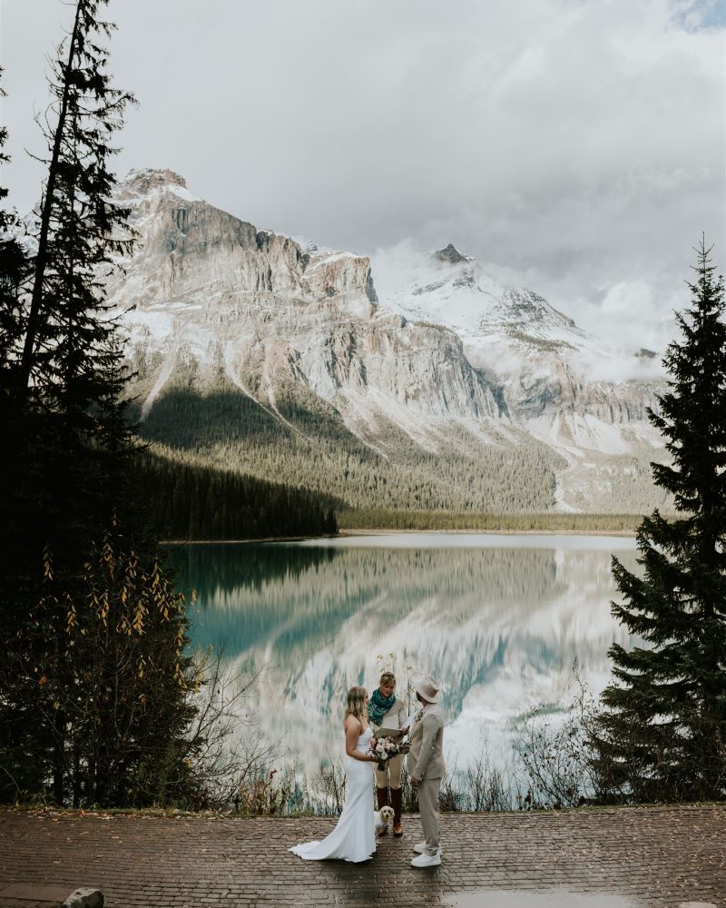 A couple stands on a stone path by a lake in Banff, exchanging elopement vows with a small child and officiant. Evergreen trees surround them as snow-capped mountains are reflected in the calm water under a partly cloudy sky, captured by their photographer.