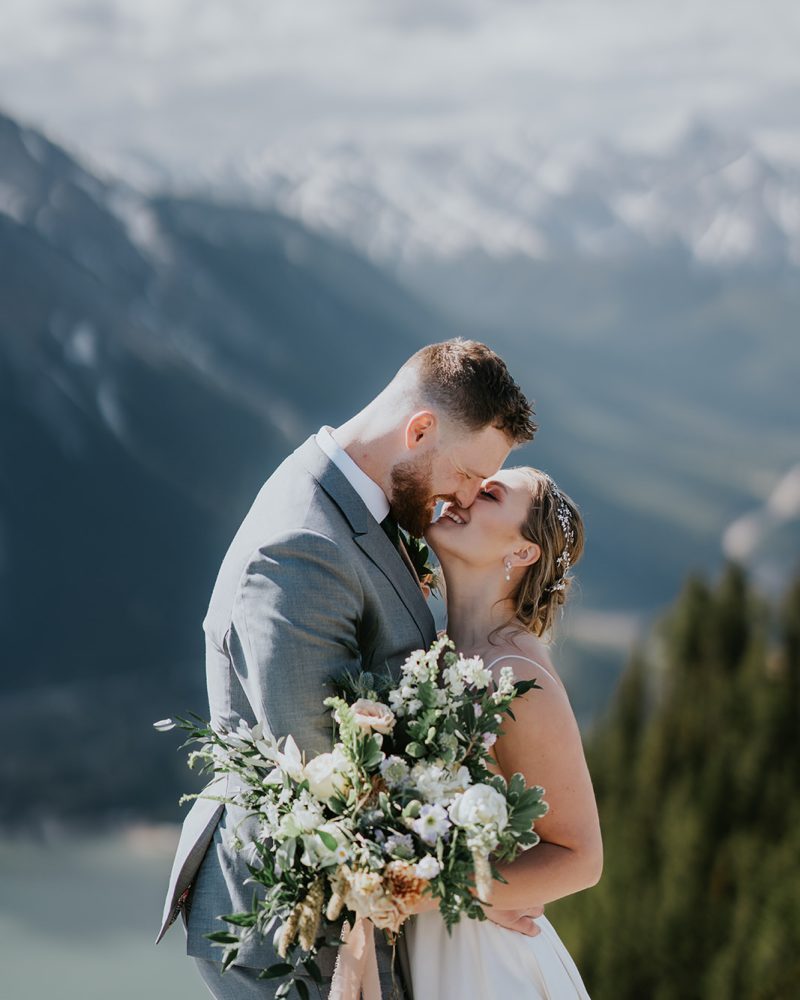 A bride and groom embrace and kiss on a mountainside in Banff, with the bride holding a bouquet of flowers. Snow-capped mountains and a lake create a breathtaking wedding backdrop.