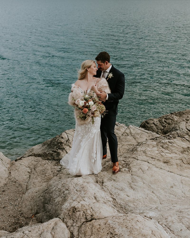 A bride and groom stand on rocky terrain by a large body of water near Canmore. The bride holds a bouquet of flowers, and the groom embraces her from behind as they look at each other affectionately on their wedding day.