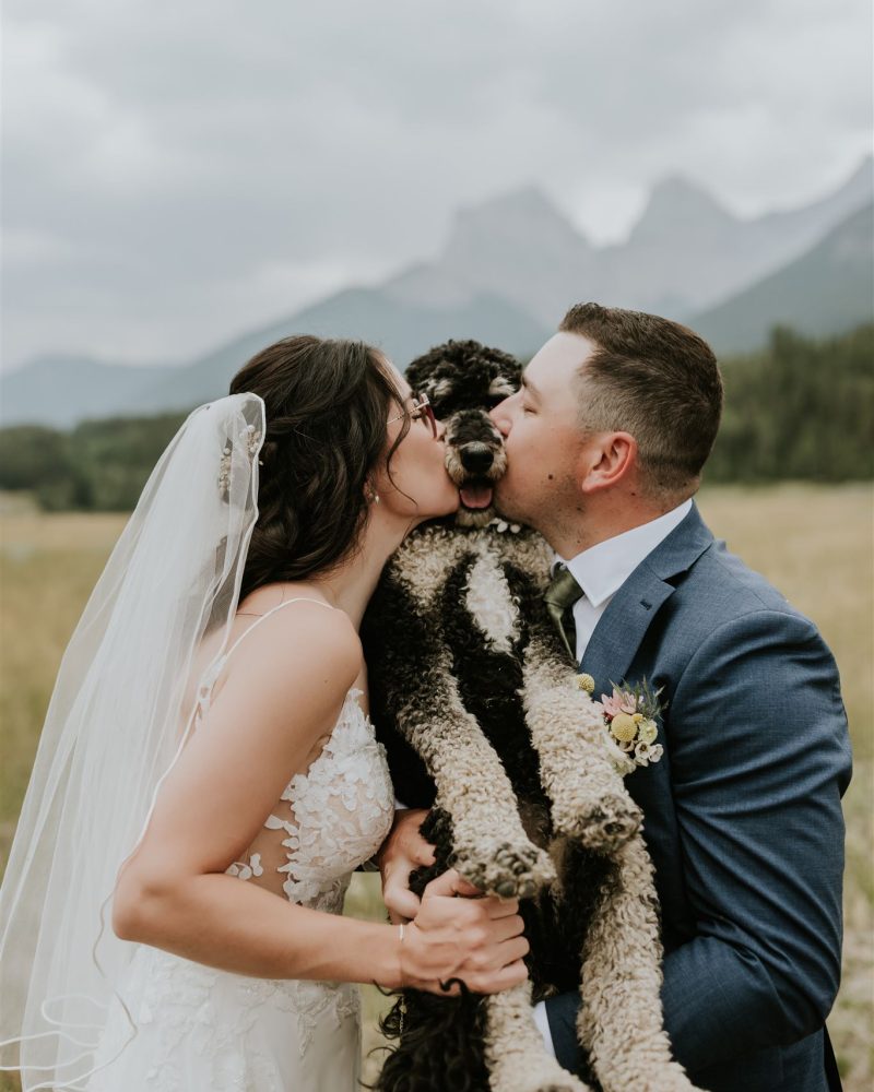A bride and groom, dressed in wedding attire, hold their black and white poodle between them and kiss its head outdoors during their Canmore elopement, with mountains and a cloudy sky in the background.