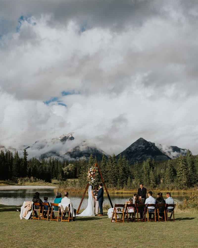 An outdoor wedding ceremony by a lake in Banff with guests seated in rows, a floral arch, and mountains covered with trees and clouds in the background under a partly cloudy sky.