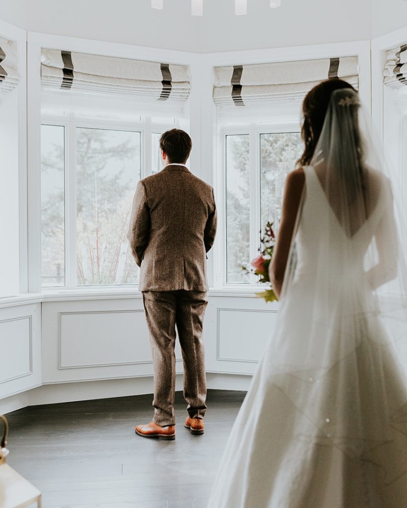 A bride in a white wedding dress approaches her groom in a brown suit, his back to her as he faces a large bay window. Captured by a Calgary photographer, their intimate elopement unfolds in a bright, elegant room.