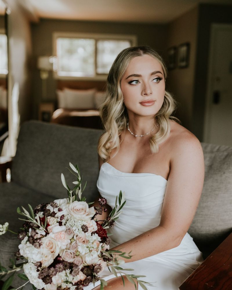 A bride in a strapless white dress sits indoors on a sofa, holding a bouquet of pale and dark blooms. She has wavy blonde hair and gazes thoughtfully to the side as natural light fills the room—captured by a Calgary photographer.