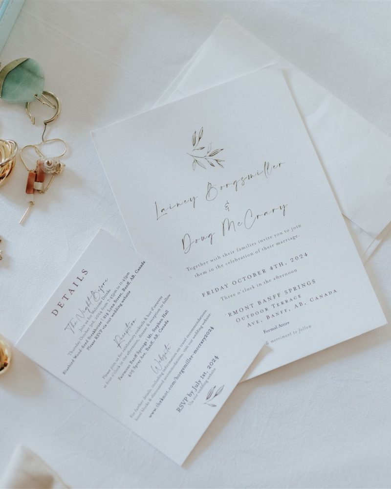 A wedding invitation and details card lie on a white surface next to gold rings, earrings, a necklace, and a blue jewelry box. The elegant, minimalist design is perfect for an intimate Canmore elopement, featuring delicate greenery artwork.