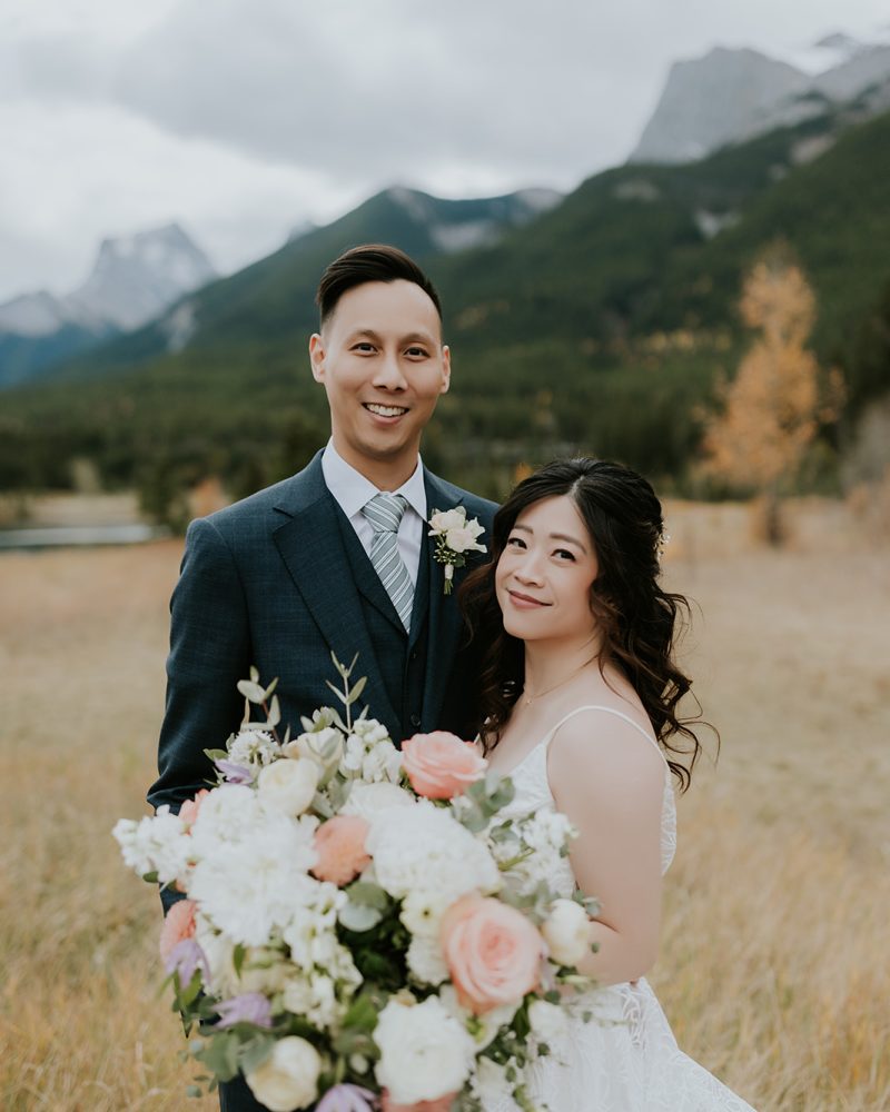A bride and groom stand outdoors in a grassy field with mountains in the background, capturing their Canmore elopement. The bride holds a large bouquet of white and pink flowers. Both are smiling at the camera on a cloudy day.