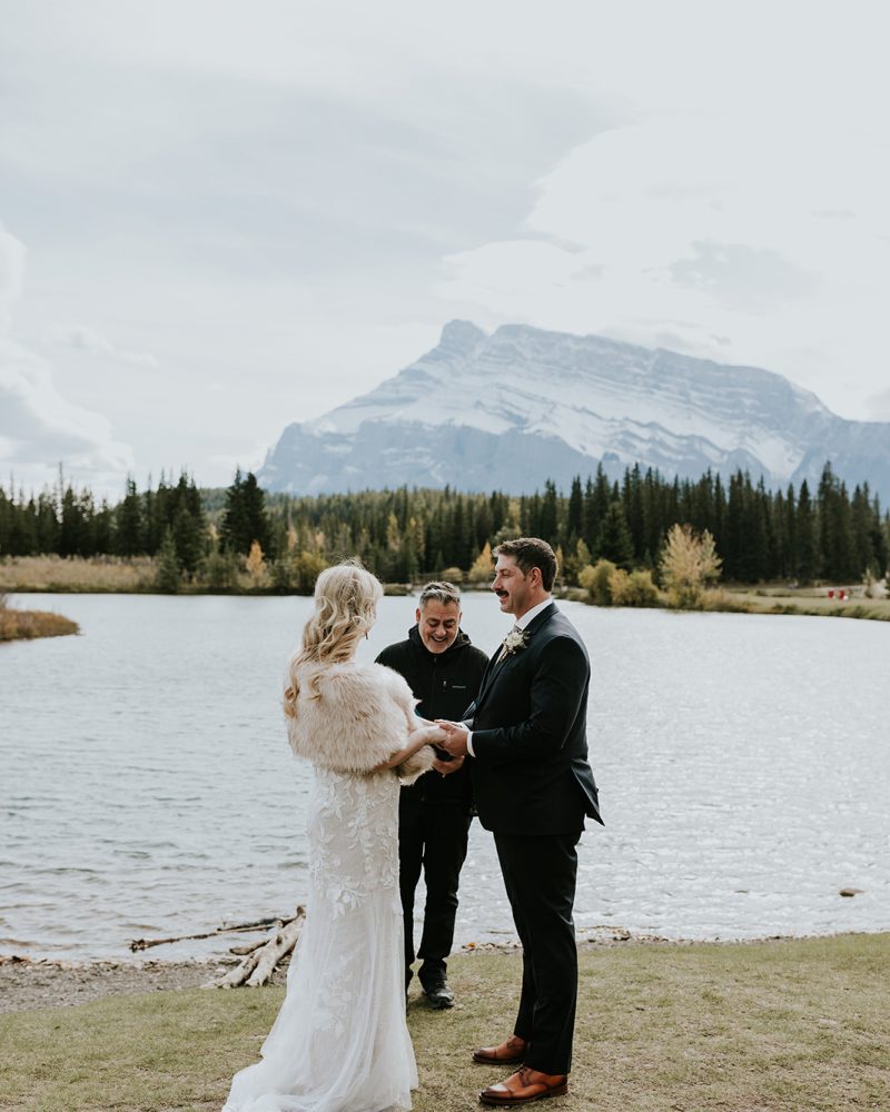 A bride and groom stand facing each other, holding hands during an outdoor wedding ceremony by a lake near Banff, with an officiant behind them and stunning mountains and pine trees in the background.