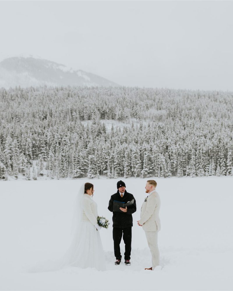 A couple in white wedding attire stands with an officiant on a snowy Canmore landscape, surrounded by snow-covered trees and mountains in the background under an overcast sky, perfectly captured by a wedding photographer.