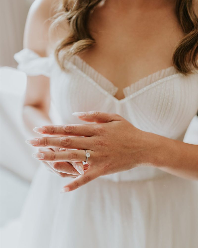 A bride in a white, off-the-shoulder wedding dress gently touches her engagement ring, showcasing delicate, manicured nails. Her wavy hair cascades over her shoulders as she stands in soft natural light during an intimate Banff elopement.