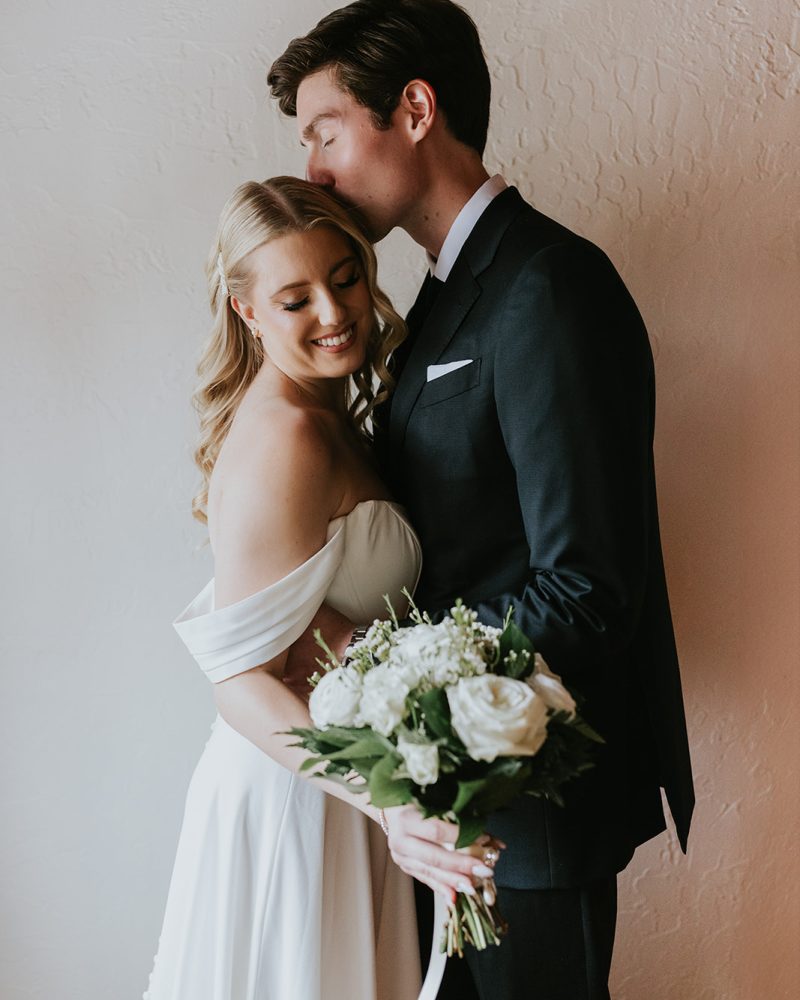 A bride in an off-shoulder white gown holds a bouquet of white flowers and smiles as a groom in a dark suit kisses her forehead—captured perfectly by their wedding photographer against a textured light wall.