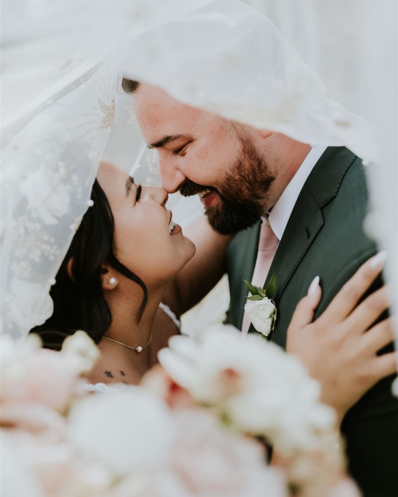 A bride and groom smile and touch noses under a sheer white veil, surrounded by soft pink and white flowers, sharing an intimate, joyful elopement moment captured by their photographer.