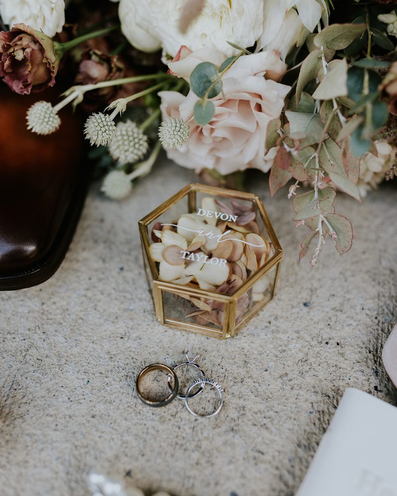 A hexagonal glass box filled with pale rose petals and labeled Devon & Taylor sits on a textured surface, surrounded by wedding rings, pearl jewelry, and a bouquet of soft-colored flowers—perfect for a romantic Banff or Canmore wedding.
