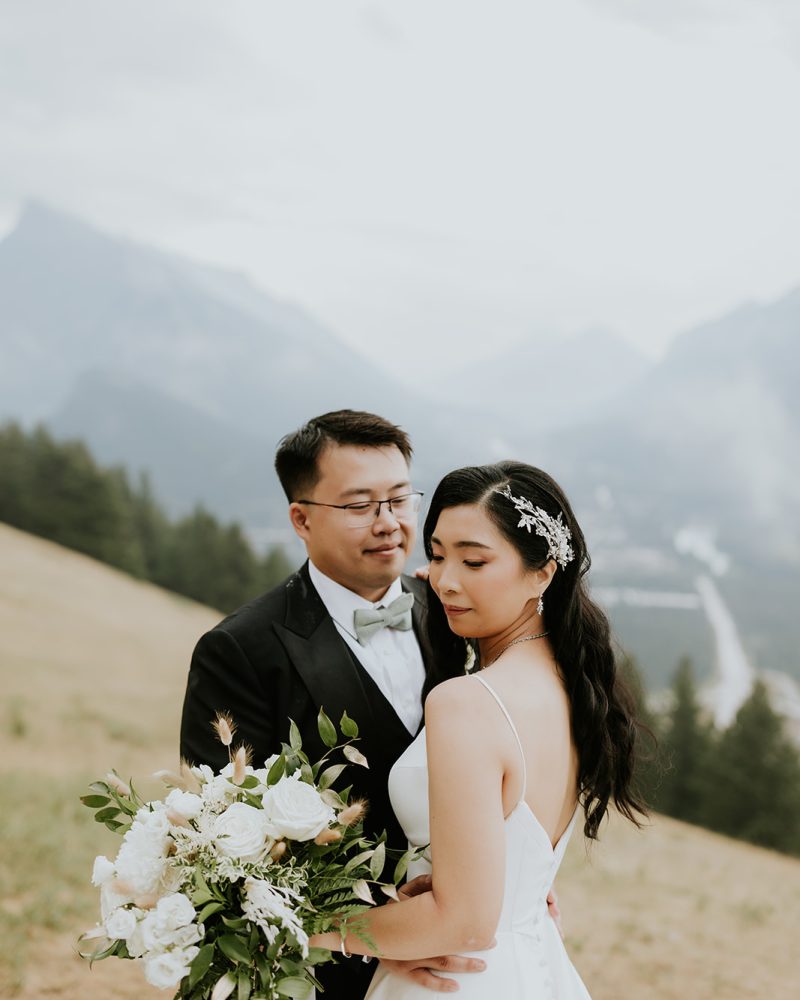 A bride and groom stand close together on a grassy Canmore hillside, surrounded by mountains and trees. Captured by their photographer, the elopement features a white bouquet, jeweled hairpiece, and the groom in a classic black suit and bow tie.