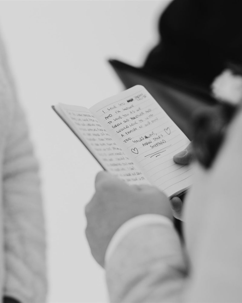 A person in a light-colored suit holds an open notebook with handwritten vows, reading aloud to someone nearby; captured in black and white by a Calgary photographer, both individuals are partially visible.
