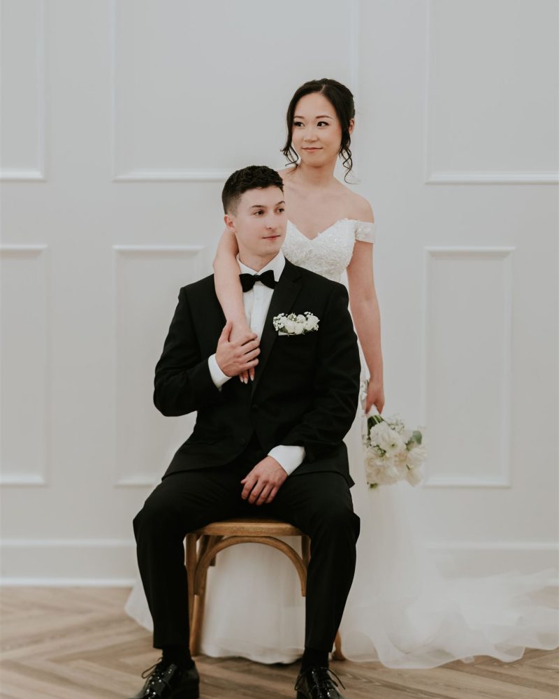 A bride in a white gown stands with her arm around a seated groom in a black tuxedo. She holds a bouquet of white flowers as the couple poses after their Calgary wedding, framed by a white paneled wall and wooden floor.
