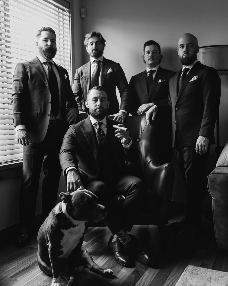 Five men in suits pose in a formal, black-and-white setting—four standing, one seated in an armchair. A dog sits on the wooden floor. Light streams through window blinds, capturing the elegance of this Banff wedding moment.