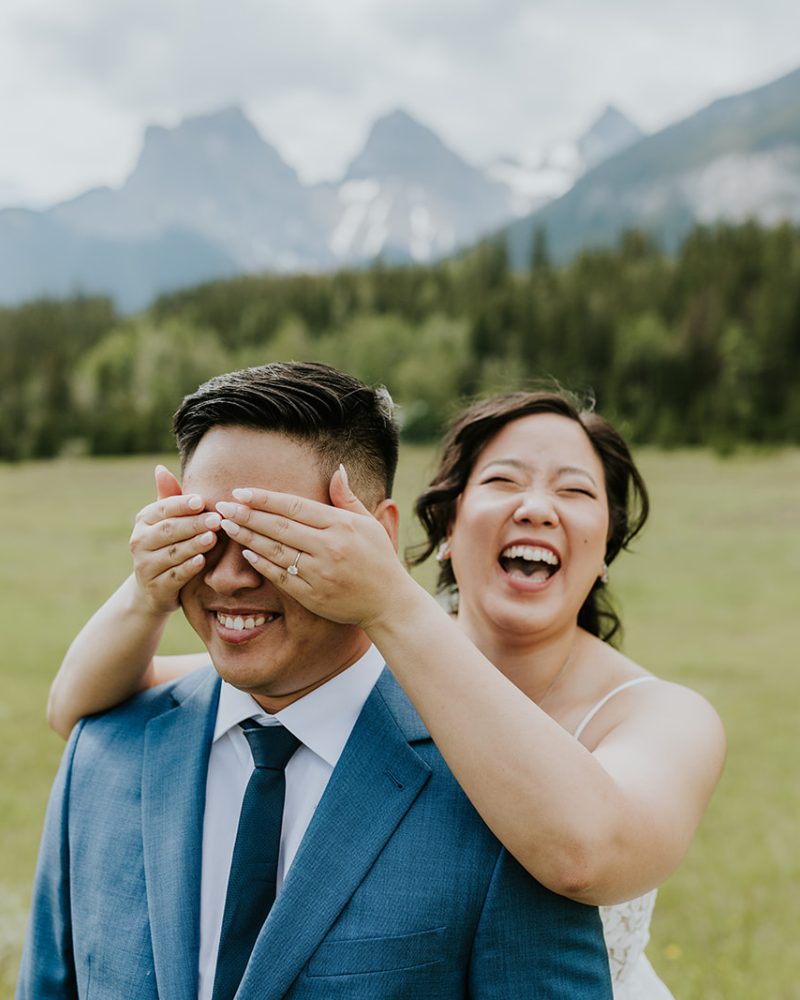 A joyful bride in a white dress playfully covers the groom's eyes from behind as he smiles, standing in a green field with mountains and trees—the perfect wedding moment captured by their photographer.