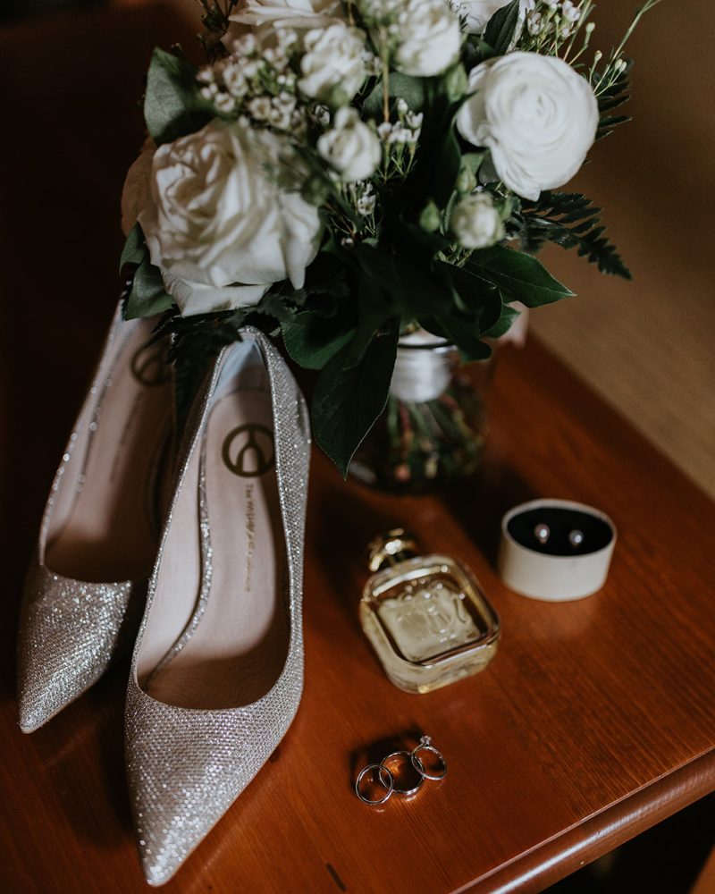 A bouquet of white roses, sparkly high-heeled shoes, a perfume bottle, and elegant rings are arranged on a wooden surface—perfect for a Calgary or Banff celebration.