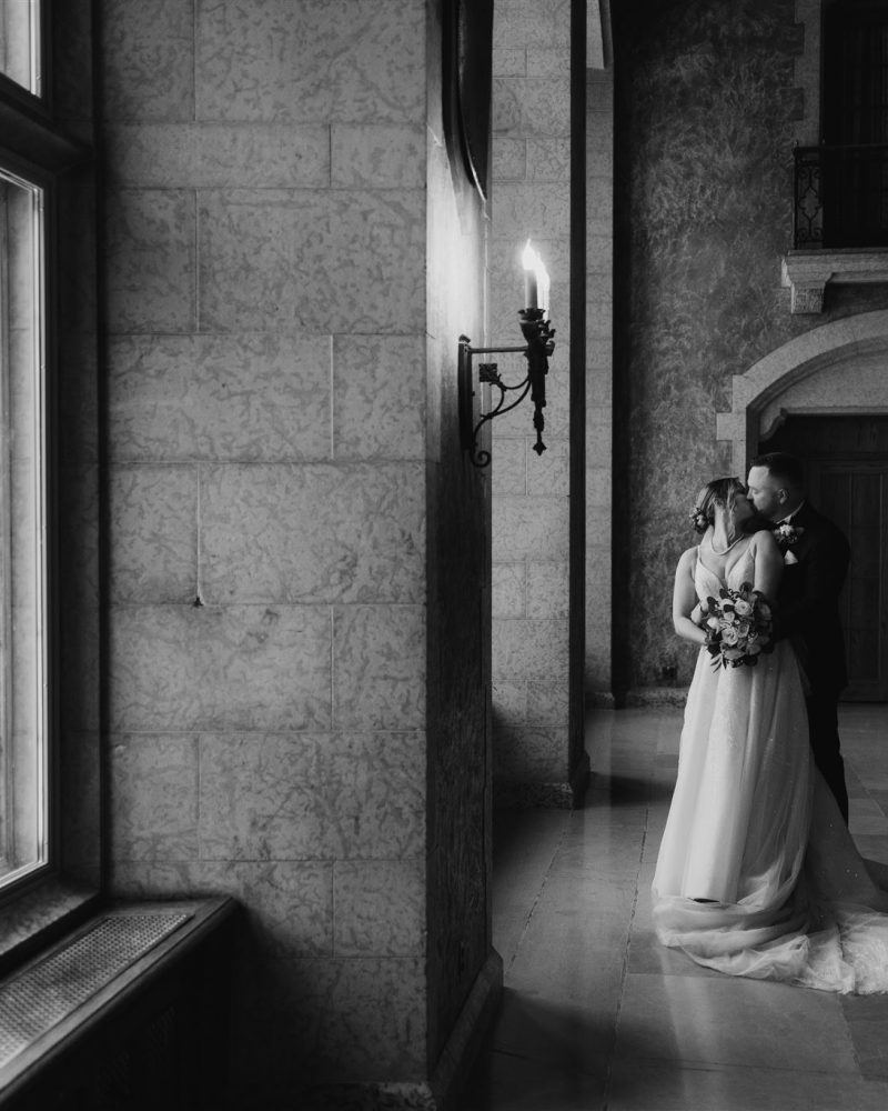 A bride and groom embrace in a grand, elegant hallway near a large window, with soft light streaming in. Capturing the intimate romance of an elopement, the black and white image evokes the timeless beauty of Banff.