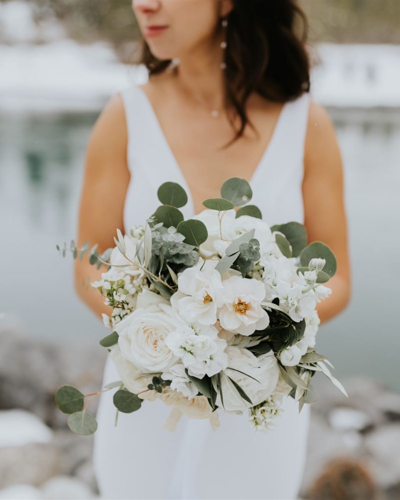 A woman in a white wedding dress holds a bouquet of white flowers and greenery, standing near a snowy lakeshore in Canmore, with water and rocks in the background. Her face is slightly out of focus.