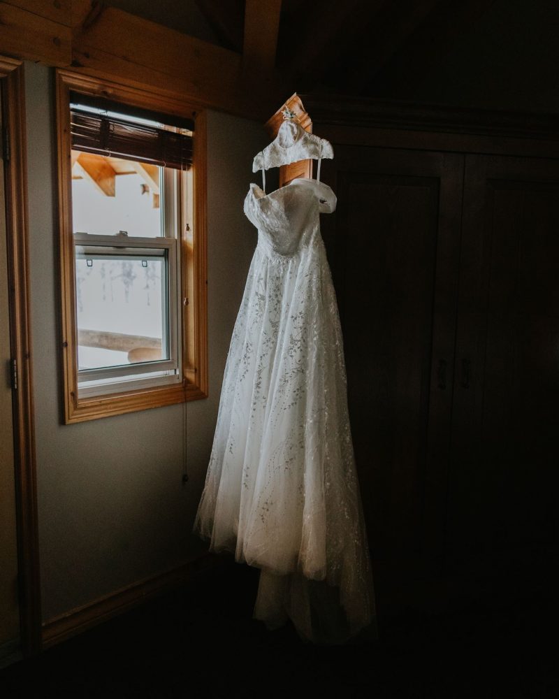 A white lace wedding dress with off-the-shoulder sleeves hangs on a wooden hanger in a dimly lit room near a window, ready for an intimate elopement in Canmore.