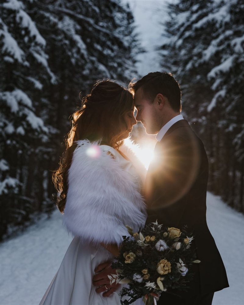 A bride and groom stand close together on a snowy Banff forest path at sunset. The bride holds a bouquet and wears a white dress with a fur shawl, while sunlight glows between their faces, creating a romantic atmosphere.