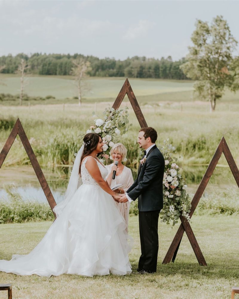 A bride and groom hold hands during an outdoor Calgary wedding ceremony, standing before wooden triangular arches adorned with flowers, surrounded by bridesmaids, groomsmen, and seated guests on a sunny day.