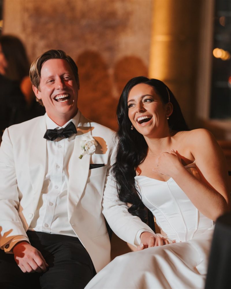 A bride and groom sit together, laughing joyfully during their Canmore wedding reception. The bride wears a white dress and the groom a white tuxedo jacket. Guests in formal attire smile in the background.
