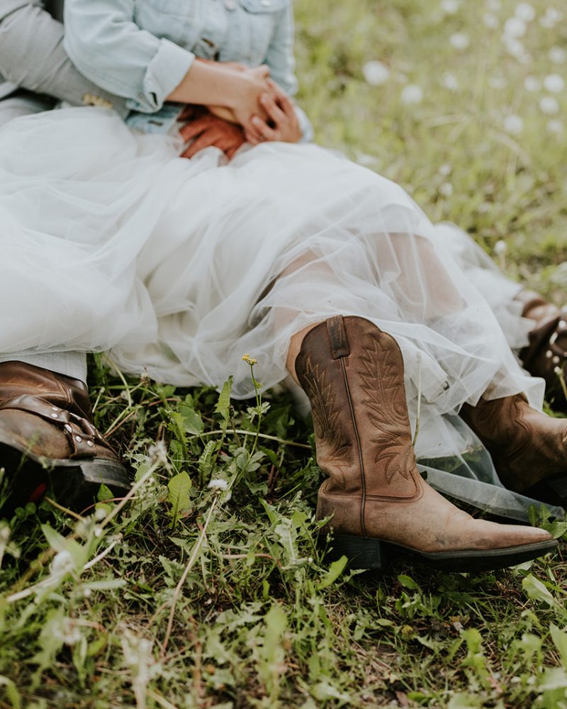 A couple sits close together in a grassy Banff field. The person in front wears a white dress and brown cowboy boots, while the person behind wraps their arms around them. Only their lower bodies are visible.