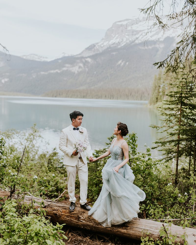 A couple dressed in formal attire stands on a fallen log by a lake in Banff, surrounded by greenery and mountains. The woman wears a flowing blue gown, the man a white suit—both holding hands and smiling at their elopement.