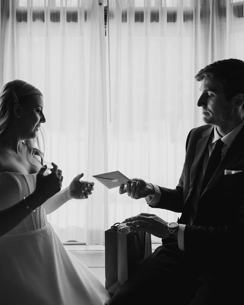 A bride and groom sit together by a window with sheer curtains, exchanging cards and gifts, both dressed formally; this intimate elopement scene is captured in black and white by a Calgary photographer.