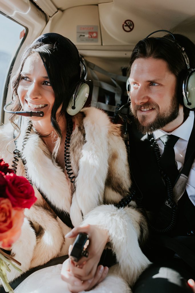 A smiling couple wearing headsets and winter coats sit inside a helicopter near Banff, holding hands. The woman holds a bouquet of red and orange flowers, sunlight streaming in—captured perfectly by their elopement photographer.
